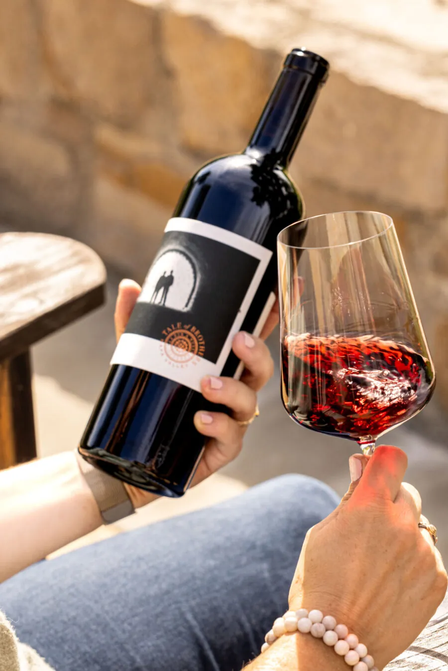 Person holding a bottle of red wine in one hand and a glass of swirling red wine in the other. Sunlight illuminates the wine and gives a warm tone to the scene. Stone wall in the background.