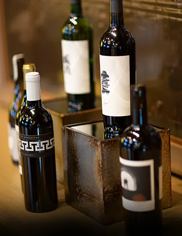 Several bottles of wine displayed on a wooden surface with one resting on a metallic stand. Some labels are detailed, while others are minimalist.