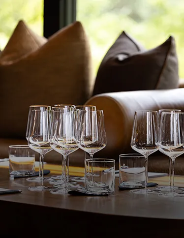 A set of empty wine glasses and tumblers arranged on a wooden table near a window with blurred cushions and pillows in the background.