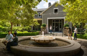 Several people are gathered around a fountain in front of a gray building with a porch and string lights, surrounded by green trees and shrubs.